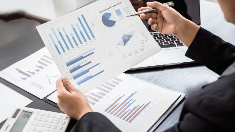 Eine Person analysiert Business Intelligence Berichte mit Diagrammen und Statistiken auf Papier, während sie Notizen auf einem Laptop macht