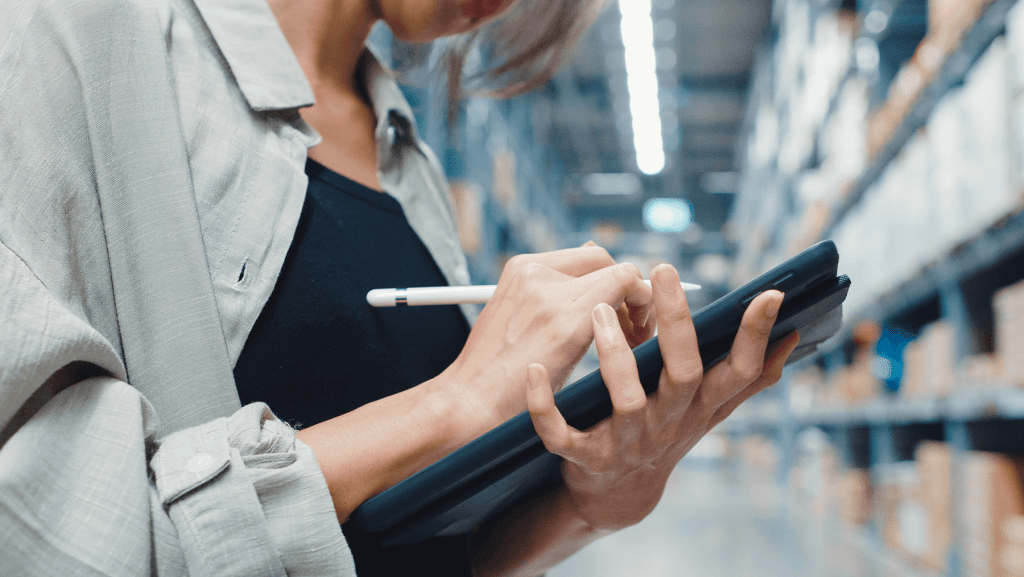 Ein Lagermitarbeiter nutzt ein Tablet im Warenlager, im Hintergrund sind Lagerregale zu sehen. Das Bild zeigt die mobile ERP-Nutzung im Arbeitsalltag.